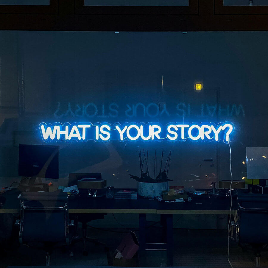 Blue Neon sign in an office window that says What Is Your Story?